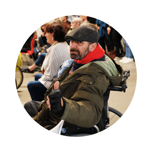 Un homme dans un fauteuil roulant portant une casquette et un manteau, faisant un geste de pouce levé en signe d'approbation, entouré de personnes dans une foule.